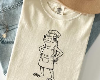 Op de afbeelding: Ivoor T-shirt met een zwart-wit illustratie van een kikkerkok met een koksmuts en schort. De tekst "needs salt" staat eronder. Het T-shirt ligt op een spijkerbroek.