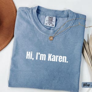 May include: Blue jean-colored t-shirt with the text "Hi, I'm Karen" in white. A gold necklace with a pendant rests on the shirt. The shirt is a Comfort Colors brand.