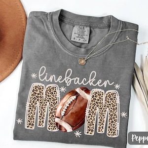 Może przedstawiać: Szara koszulka z białym napisem "linebacker mom" i grafiką piłki do futbolu amerykańskiego. Litery "M" mają wzór w panterkę. Na koszulce znajduje się srebrny naszyjnik z wisiorkiem.