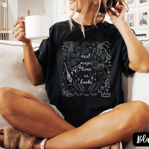 Puede incluir: Camiseta negra con un dise&ntilde;o gr&aacute;fico blanco que presenta dragones, alas, estrellas y el texto "real magic lives in books". La camiseta es de estilo c&oacute;modo e informal.