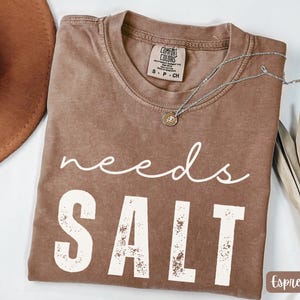 Puede incluir: Camiseta color espresso con las palabras blancas "needs SALT". La camiseta tiene cuello redondo y una etiqueta Comfort Colors. Un collar plateado con un pequeño colgante descansa sobre la camiseta. Un sombrero marrón es visible en la esquina superior izquierda.