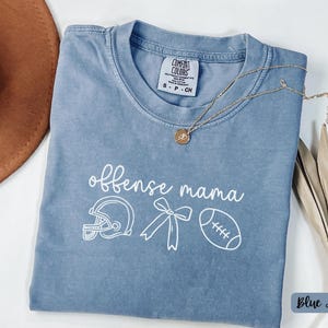 Könnte beinhalten: Blaues Jeans-T-Shirt mit den Worten "offense mama" und Football-Grafiken. Das Shirt zeigt einen Footballhelm, eine Schleife und einen Football. Eine goldene Halskette ist über dem Shirt drapiert.
