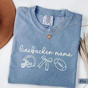 Può includere: T-shirt blu jeans con le parole "linebacker mama" e grafica a tema football americano. La maglietta ha girocollo e maniche corte. Una collana d'oro è drappeggiata sulla maglietta.