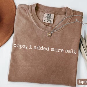 Puede incluir: Camiseta color espresso con la frase "oops, i added more salt" en letras blancas. Un collar plateado con un pequeño colgante descansa sobre la camisa. La camiseta tiene una etiqueta Comfort Colors.