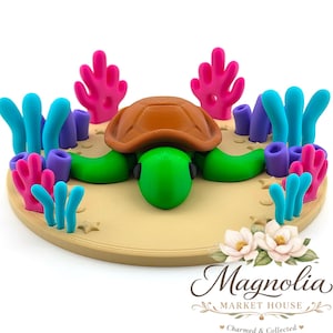 May include: A handcrafted, colorful turtle figurine with a brown shell, green head and limbs, surrounded by coral and seaweed. The turtle is set on a tan base with a floral logo in the bottom right corner. The coral is pink, and the seaweed is blue and purple.
