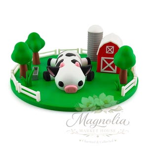 May include: A miniature farm scene featuring a black and white cow resting on green grass within a white picket fence. The scene includes a red barn, a silo, and green trees. The base is a green circle.