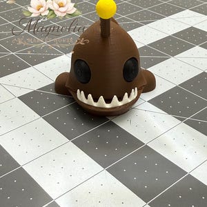 May include: A brown 3D printed creature figurine with a smooth, rounded head, two black circular eyes, and a wide mouth filled with sharp white teeth. A thin stalk extends from the top of the head, topped with a small, bright yellow sphere.