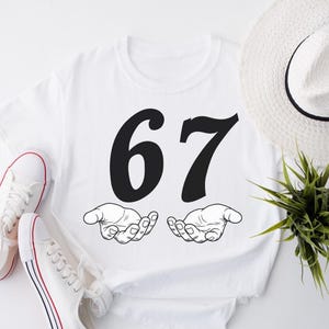 May include: White t-shirt with the number 67 in black, above two open hands. A pair of white canvas sneakers with red and blue stripes and a white hat with a black band are also in the image. A small green plant is also visible.