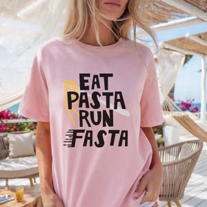 Peut inclure: T-shirt rose clair avec l'inscription "EAT PASTA RUN FASTA" en noir. Le design comprend une fourchette à pâtes jaune et un brin de pâtes blanc. Le t-shirt est de style décontracté, à manches courtes.