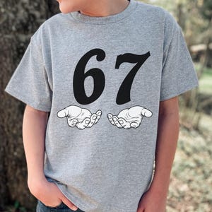 May include: A gray t-shirt featuring the number "67" in bold black font above two stylized open hands. The shirt is made of a soft material and is suitable for casual wear.