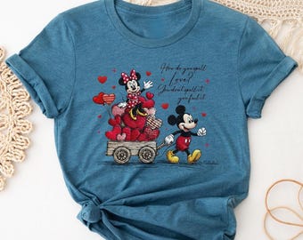 May include: Blue t-shirt featuring Mickey Mouse pulling a cart filled with heart-shaped balloons and Minnie Mouse. The shirt has the text "How do you spell love? You don't spell it, you feel it."