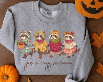 Pode incluir: Um moletom cinza com uma ilustração lúdica de quatro guaxinins em trajes de outono, de mãos dadas. Os guaxinins usam casacos, chapéus e suéteres. O texto "just a cozy raccoon" está impresso abaixo. Uma abóbora esculpida e uma abóbora pequena estão na imagem.