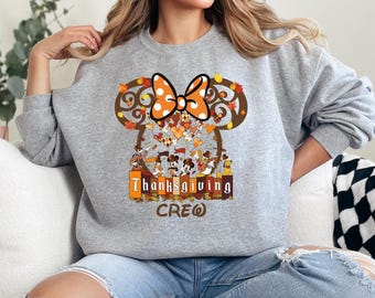 May include: A gray sweatshirt featuring a Thanksgiving design. The design includes a Minnie Mouse bow with fall leaves, a collection of cartoon characters, and the words "Thanksgiving Crew".