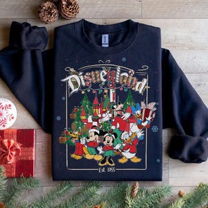 K&ouml;nnte beinhalten: Marineblaues Disneyland-Sweatshirt mit einer farbenfrohen Grafik mit Micky Maus, Minnie Maus, Donald Duck und anderen Disney-Figuren. Die Grafik enth&auml;lt den Text "Disneyland" und "Est. 1955". Festliches Weihnachtsmotiv.
