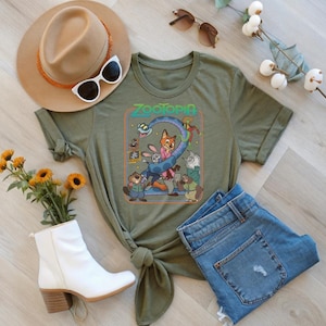 May include: Olive green t-shirt featuring the word "Zootopia" and cartoon characters. The shirt is styled with blue jeans, a tan hat, white sunglasses, and a pair of white ankle boots. A bouquet of yellow flowers and a sprig of white flowers are also included.