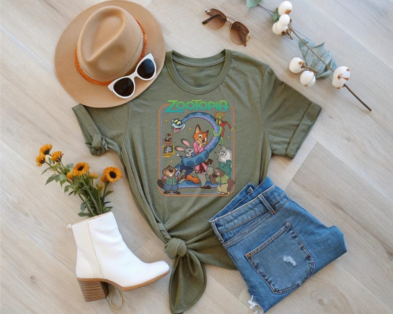 May include: Olive green t-shirt featuring the word "Zootopia" and cartoon characters. The shirt is styled with blue jeans, a tan hat, white sunglasses, and a pair of white ankle boots. A bouquet of yellow flowers and a sprig of white flowers are also included.
