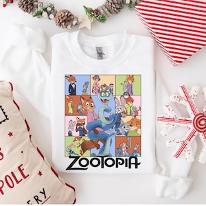 Pu&ograve; includere: Felpa bianca con una grafica colorata dei personaggi del film d'animazione Zootopia. Il design &egrave; diviso in pannelli rettangolari con la scritta "Zootopia" in lettere nere. Decorazioni natalizie circondano la felpa.