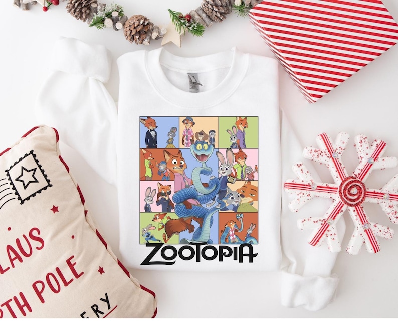 Pu&ograve; includere: Felpa bianca con una grafica colorata dei personaggi del film d'animazione Zootopia. Il design &egrave; diviso in pannelli rettangolari con la scritta "Zootopia" in lettere nere. Decorazioni natalizie circondano la felpa.