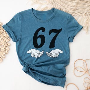 May include: A blue t-shirt with the number 67 in black, above two cartoon hands. The hands are white with black outlines, palms up. The shirt is folded at the bottom.