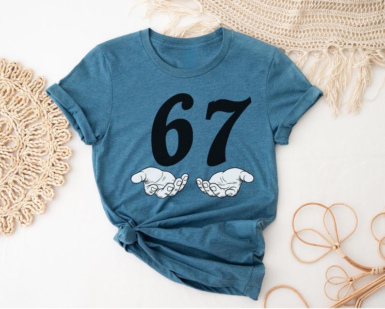 May include: A blue t-shirt with the number 67 in black, above two cartoon hands. The hands are white with black outlines, palms up. The shirt is folded at the bottom.
