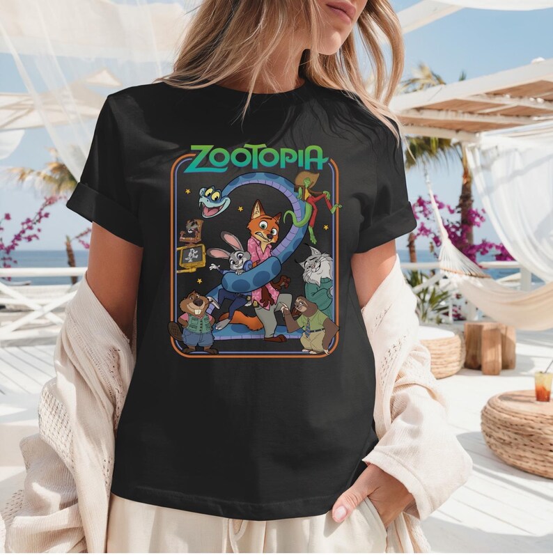 May include: Black t-shirt featuring a colorful graphic from the animated film "Zootopia." The design includes the film's title and various characters, such as Judy Hopps and Nick Wilde, in a retro-style frame. The shirt has short sleeves.