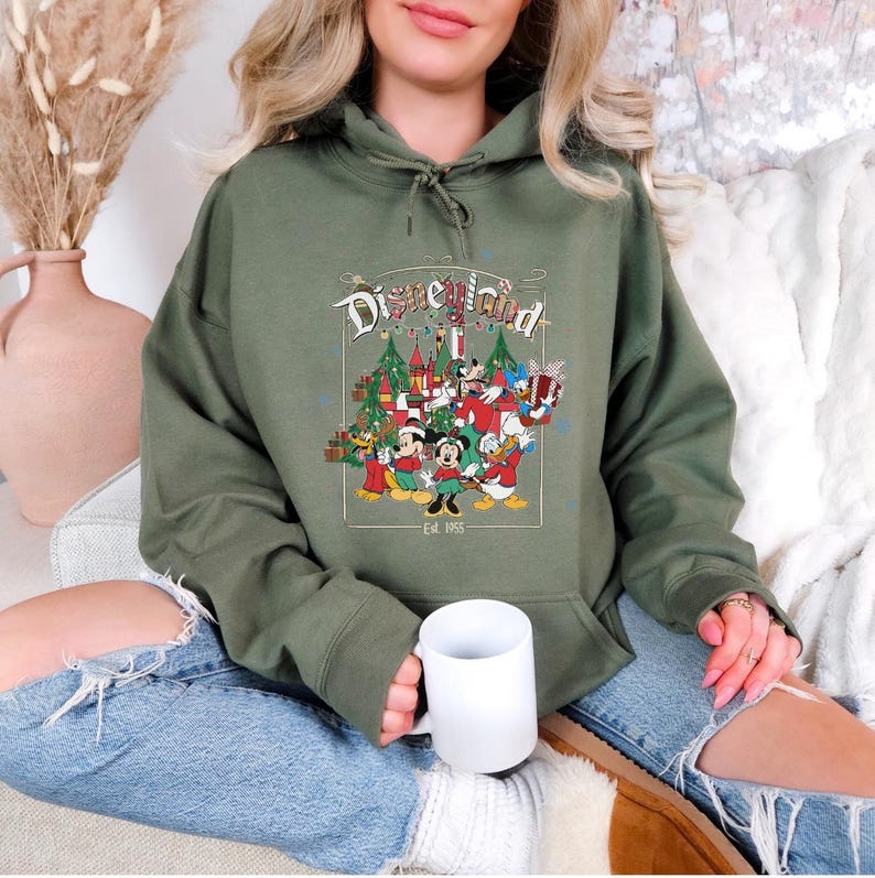 K&ouml;nnte beinhalten: Ein olivgr&uuml;ner Kapuzenpullover mit einem Disneyland-Motiv mit Micky Maus, Minnie Maus, Donald Duck und anderen Figuren. Die Grafik enth&auml;lt Weihnachtsb&auml;ume und ein Schloss. Der Text "Disneyland" und "Est. 1955" sind ebenfalls sichtbar.