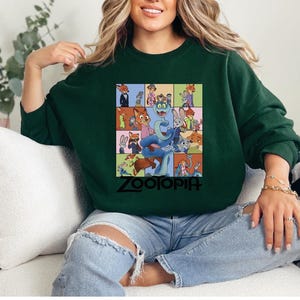 Pu&ograve; includere: Una felpa verde scuro con una grafica colorata dei personaggi del film d'animazione "Zootropolis". La grafica &egrave; una griglia di immagini con la parola "Zootopia" in nero in basso. La felpa &egrave; indossata da una persona.