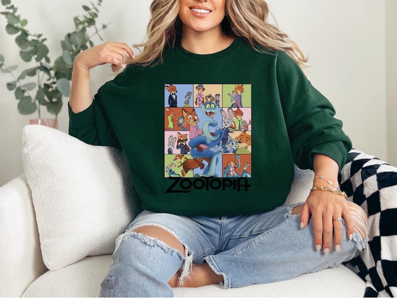Pu&ograve; includere: Una felpa verde scuro con una grafica colorata dei personaggi del film d'animazione "Zootropolis". La grafica &egrave; una griglia di immagini con la parola "Zootopia" in nero in basso. La felpa &egrave; indossata da una persona.