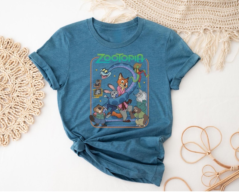 May include: Teal blue t-shirt featuring a graphic from the animated movie Zootopia. The graphic includes the word "Zootopia" and several cartoon characters. The shirt has rolled-up sleeves and a tied knot at the bottom.
