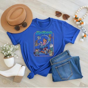 May include: A royal blue t-shirt featuring a graphic from the movie "Zootopia". The design includes characters like Nick Wilde, Judy Hopps, and others. The shirt is styled with a brown hat, sunglasses, and blue jeans.