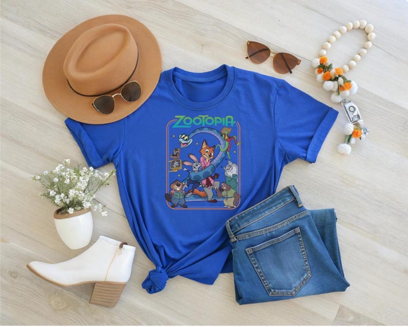 May include: A royal blue t-shirt featuring a graphic from the movie "Zootopia". The design includes characters like Nick Wilde, Judy Hopps, and others. The shirt is styled with a brown hat, sunglasses, and blue jeans.