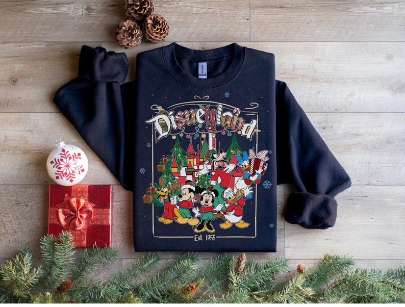 K&ouml;nnte beinhalten: Marineblaues Disneyland-Sweatshirt mit einer farbenfrohen Grafik mit Micky Maus, Minnie Maus, Donald Duck und anderen Disney-Figuren. Die Grafik enth&auml;lt den Text "Disneyland" und "Est. 1955". Festliches Weihnachtsmotiv.
