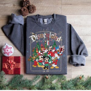 K&ouml;nnte beinhalten: Ein dunkelgraues Disneyland-Sweatshirt mit einer farbenfrohen Grafik mit Micky Maus, Minnie Maus, Donald Duck und Goofy. Die Grafik enth&auml;lt Weihnachtsb&auml;ume und den Text "Disneyland" und "Est. 1955".