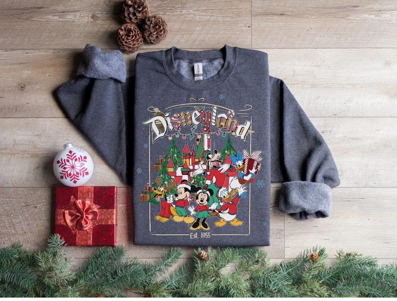 K&ouml;nnte beinhalten: Ein dunkelgraues Disneyland-Sweatshirt mit einer farbenfrohen Grafik mit Micky Maus, Minnie Maus, Donald Duck und Goofy. Die Grafik enth&auml;lt Weihnachtsb&auml;ume und den Text "Disneyland" und "Est. 1955".