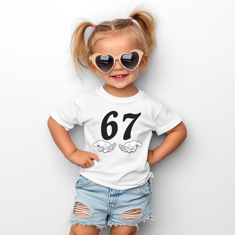 May include: A young child wearing a white t-shirt with the number "67" printed in black, along with two open hands. The child is also wearing heart-shaped sunglasses and denim shorts.
