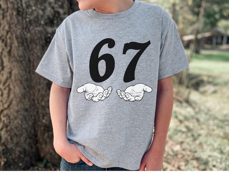 May include: A gray t-shirt featuring the number "67" in bold black font above two stylized open hands. The shirt is made of a soft material and is suitable for casual wear.