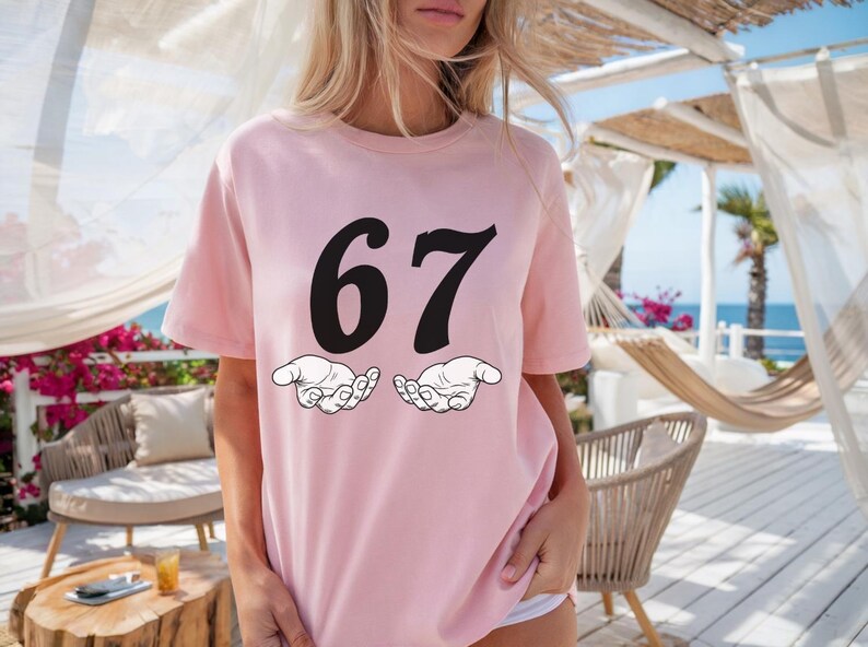 May include: A light pink t-shirt with the number "67" in bold black font. Below the numbers are two white cartoon hands. The shirt is a casual style.