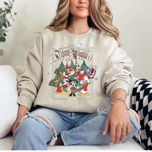 K&ouml;nnte beinhalten: Beiges Disneyland-Sweatshirt mit einer farbenfrohen Grafik von Disney-Figuren und einem Schloss, mit dem Text "Disneyland". Das Design umfasst Weihnachtsb&auml;ume und Schneeflocken und schafft ein festliches Urlaubsthema.
