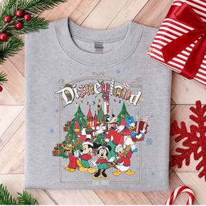 K&ouml;nnte beinhalten: Graues Sweatshirt mit Rundhalsausschnitt und Disneyland-Grafik. Die Grafik zeigt Mickey Mouse, Minnie Mouse, Donald Duck und Goofy in festlicher Kleidung, ein Schloss, Weihnachtsb&auml;ume und den Text "Disneyland" und "Est. 1955".