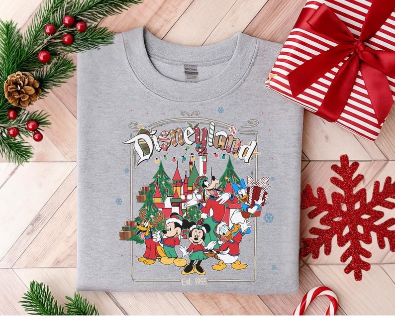 K&ouml;nnte beinhalten: Graues Sweatshirt mit Rundhalsausschnitt und Disneyland-Grafik. Die Grafik zeigt Mickey Mouse, Minnie Mouse, Donald Duck und Goofy in festlicher Kleidung, ein Schloss, Weihnachtsb&auml;ume und den Text "Disneyland" und "Est. 1955".