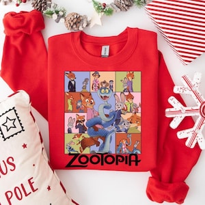 Pu&ograve; includere: Una felpa girocollo rossa con una grafica colorata dei personaggi del film "Zootropolis". La parola "Zootopia" &egrave; stampata in nero in basso. La felpa &egrave; circondata da decorazioni natalizie, tra cui un cuscino, una confezione regalo e un ornamento a forma di fiocco di neve.