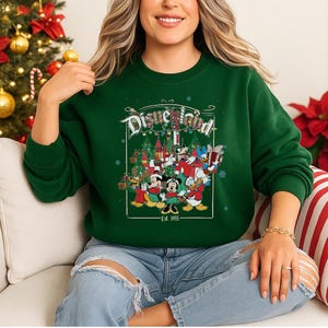 K&ouml;nnte beinhalten: Ein gr&uuml;nes Sweatshirt mit Rundhalsausschnitt und einem Disneyland-Motiv mit Mickey Mouse, Minnie Mouse, Donald Duck und anderen Figuren in einer Weihnachtsszene. Der Aufdruck enth&auml;lt den Text "Disneyland" und "Est. 1955".