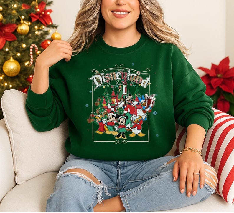 K&ouml;nnte beinhalten: Ein gr&uuml;nes Sweatshirt mit Rundhalsausschnitt und einem Disneyland-Motiv mit Mickey Mouse, Minnie Mouse, Donald Duck und anderen Figuren in einer Weihnachtsszene. Der Aufdruck enth&auml;lt den Text "Disneyland" und "Est. 1955".