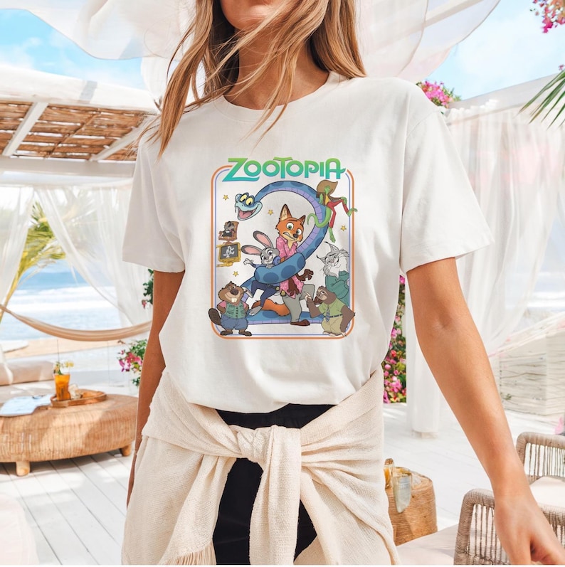 May include: White t-shirt featuring the word "Zootopia" in teal and a colorful graphic of characters from the animated movie. The design includes a fox, rabbit, and other animal characters. The shirt is a casual, short-sleeved style.