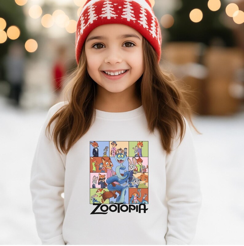 Pu&ograve; includere: Una persona giovane indossa una felpa bianca con una grafica colorata dei personaggi del film "Zootropolis". La felpa ha la scritta "Zootopia" stampata in nero. La persona indossa anche un cappello invernale rosso e bianco.