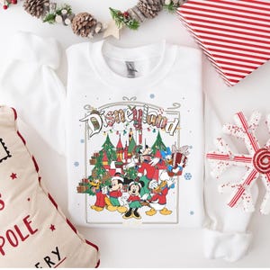 K&ouml;nnte beinhalten: Wei&szlig;es Sweatshirt mit Rundhalsausschnitt und farbenfrohem Disneyland-Motiv mit Mickey Mouse, Minnie Mouse, Donald Duck und Goofy vor einem Schloss und Weihnachtsb&auml;umen. Das Sweatshirt ist von Weihnachtsdekorationen umgeben.