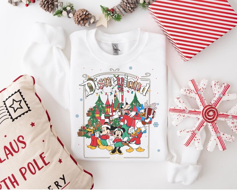 K&ouml;nnte beinhalten: Wei&szlig;es Sweatshirt mit Rundhalsausschnitt und farbenfrohem Disneyland-Motiv mit Mickey Mouse, Minnie Mouse, Donald Duck und Goofy vor einem Schloss und Weihnachtsb&auml;umen. Das Sweatshirt ist von Weihnachtsdekorationen umgeben.