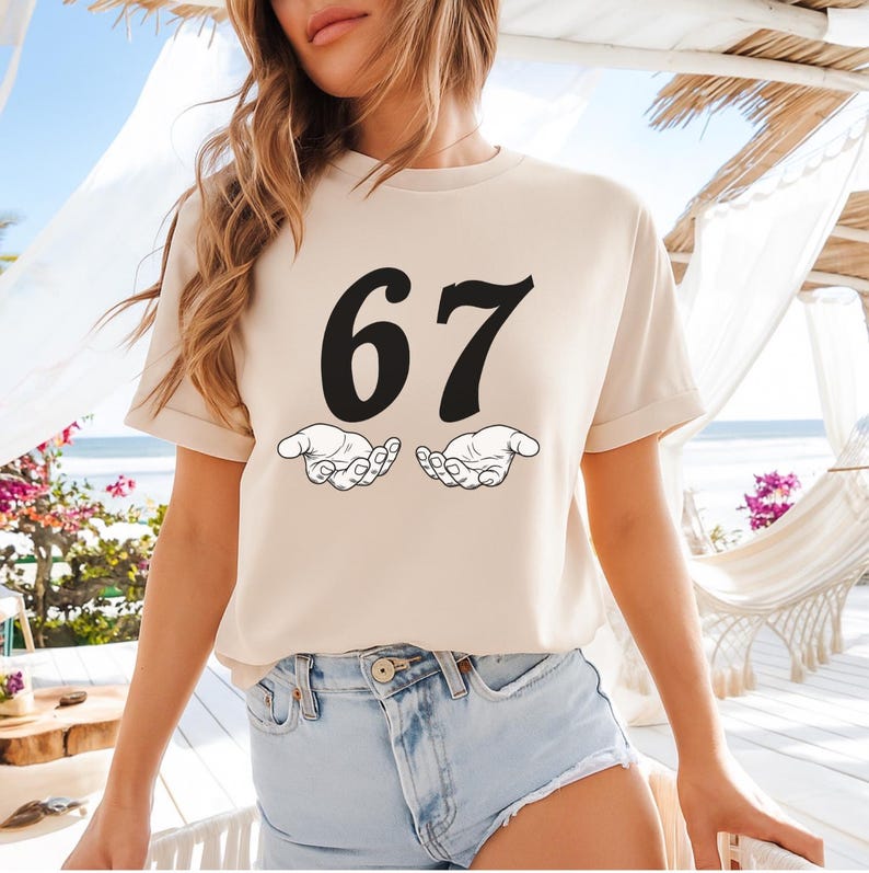 May include: A beige t-shirt featuring the number "67" in bold black font, with two open hands below. The shirt has short sleeves and a crew neck. The overall design is simple and graphic.