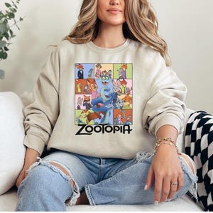 Pu&ograve; includere: Felpa beige con una grafica colorata dei personaggi del film d'animazione "Zootropolis". Il design presenta vari personaggi animali in un motivo a griglia, con la parola "Zootopia" in lettere nere in basso.