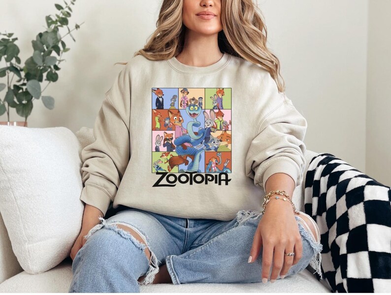 Pu&ograve; includere: Felpa beige con una grafica colorata dei personaggi del film d'animazione "Zootropolis". Il design presenta vari personaggi animali in un motivo a griglia, con la parola "Zootopia" in lettere nere in basso.